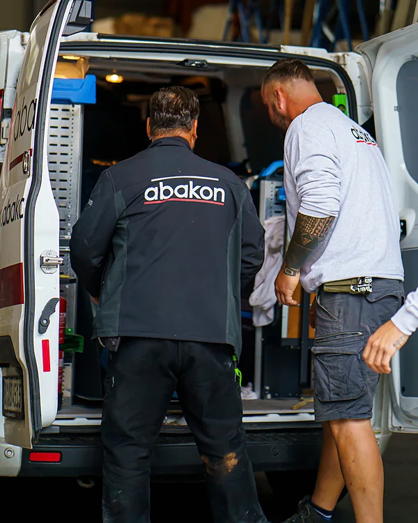 Zwei Mitarbeiter am geöffneten Transporter in der Halle. Einer trägt eine Jacke mit abakon-Logo.