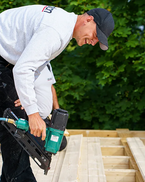 Ein junger Mitarbeiter arbeitet lachend mit einem Druckluftnagler auf einer Holzschalung.